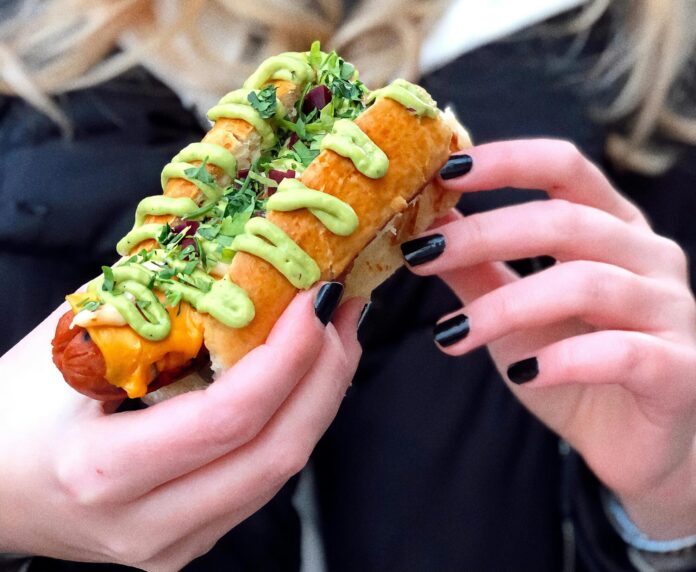 Close-Up Photo of a Woman with Black Manicured Fingernails Holding a Delicious Hot Dog Sandwich