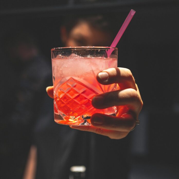 Man Holding Forward A Glass Of Iced Pinkish Colored Cocktail With A Straw
